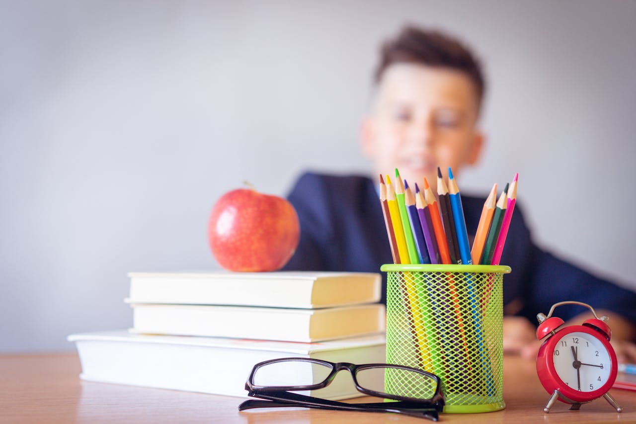 brand-01 Schoolboy smiling behind a desk with books, pencils, and an alarm clock symbolizing study and creativity.